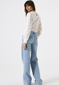 Mujer con el cabello largo y castaño, vistiendo una blusa blanca de encaje de mangas largas y unos pantalones vaqueros de tiro alto y pernera ancha en azul claro, mirando por encima del hombro.