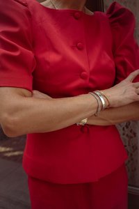 Blusa rossa su misura con maniche a palloncino e dettagli con bottoni, abbinata a una gonna coordinata. Braccialetti multipli adornano i polsi.