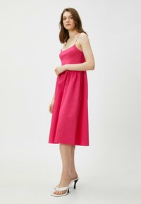 Mekko, jossa on fuksia väri, spagettiolkaimet, istuva yläosa ja runsas hame. Yhdistetty valkoisiin korkokenkiin neutraalilla taustalla.