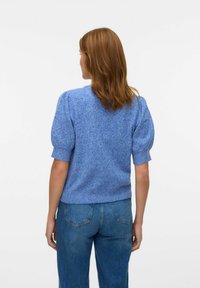 Pull en maille bleu avec des manches bouffantes courtes et une texture tachetée de lilas. Porté avec un jean en denim taille haute, vu de dos.