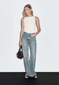 Top senza maniche di un beige chiaro con dettaglio a bottoni, abbinato a jeans a campana di un lavaggio chiaro. Il modello tiene una piccola borsa a tracolla di colore marrone scuro.