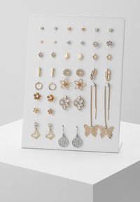 Ett örhänge-set utställt på en vit ställning, med guld- och silvertoner samt blommiga, geometriska och fjärilsdesigner. Olika texturer och former.