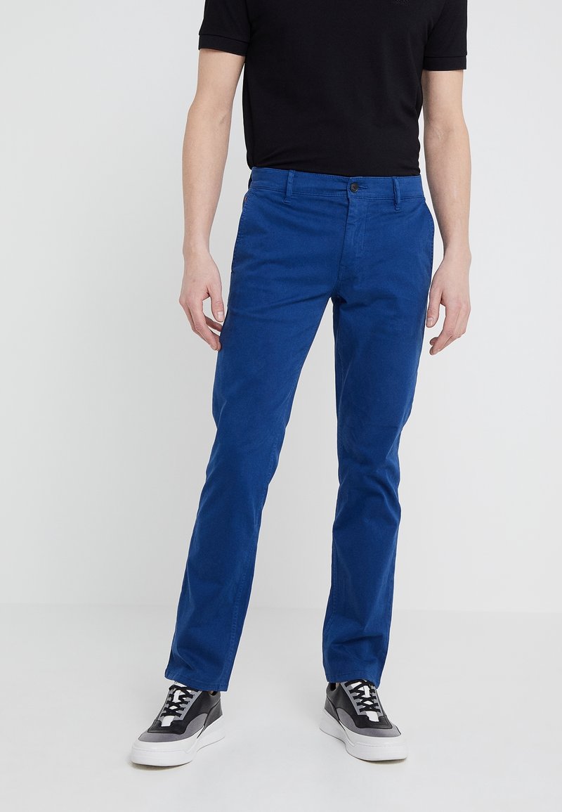 Homme portant un pantalon bleu vif associé à des baskets noires et blanches et une chemise noire à manches courtes, debout devant un fond uni.