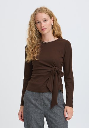 Femme aux cheveux blonds bouclés portant un haut à manches longues marron foncé noué à la taille et un pantalon gris devant un fond clair uni.