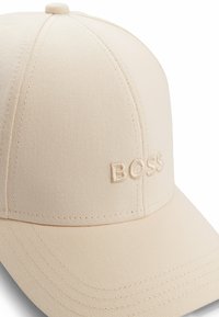 Beige baseball cap with embroidered "BOSS" logo. Made of structured fabric, features a rounded crown and pre-curved visor.
