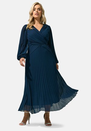 Vrouw draagt een navy blauwe, geplooide maxi-jurk met lange mouwen en een V-hals met zilveren sandalen met hoge hakken, staand tegen een effen achtergrond.