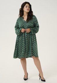 Robe verte avec un décolleté en V, des manches longues et une silhouette fluide. Présente des motifs de feuilles d'or et un tissu texturé. Chaussures à talons hauts noires.