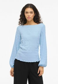 Donna con capelli ricci che indossa un top a maniche lunghe azzurro chiaro con tessuto strutturato e pantaloni neri a gamba larga, in piedi su uno sfondo bianco.