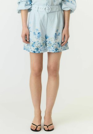 Vrouw draagt lichtblauwe korte broek met bloemmotief en bijpassende riem en zwarte sandalen, staand tegen een effen witte achtergrond.