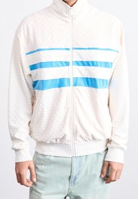 Man i vit texturerad zip-up jacka med tre horisontella blå och vita ränder över bröstet samt ljusblå jeans.