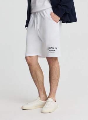 Uomo che indossa pantaloncini bianchi con coulisse con la scritta "Authentic Co", sneakers bianche e una giacca blu navy con le mani nelle tasche.
