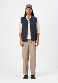 Marineblauw gewatteerd vest met hexagonaal patroon, beige trui, tan kleurige broek en zwarte schoenen. Model staat tegen een effen witte achtergrond.