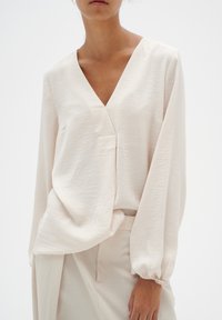 Hellbeige Bluse mit V-Ausschnitt, langen puffärmel und strukturiertem Stoff. Sanfte, fließende Form mit dezenten Nähdetails.