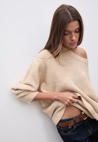 Bežna oversized jopica s širokimi rokavi, izdelana iz mehkega pletenega materiala; nosi se čez temne kavbojke, ki jih dopolnjuje rjav pas z diskretnimi kovinskimi elementi.