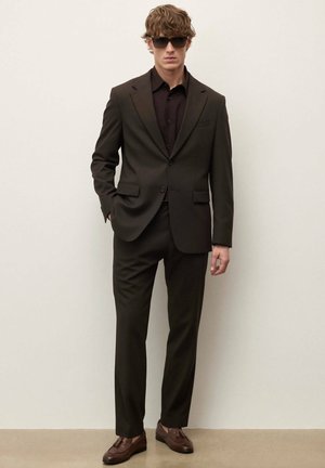 Mlad moški v temno rjavi obleki, črni srajci, rjavih usnjenih mokasinah s cofki in velikih črnih sončnih očalih, ki stoji ob svetlem enobarvnem ozadju.