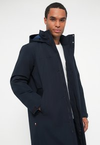 Mann in einem navyblauen Wintermantel mit Kapuze, Reißverschluss und Druckknöpfen, der mit einer Hand in der Tasche gegen einen schlichten Hintergrund posiert.