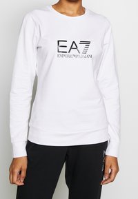 Vit sweatshirt i bomull med långa ärmar. Har en reflekterande svart logotyp "EA7 Emporio Armani" på framsidan och ribbade muddarmar.