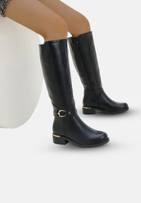 Bottes en cuir noir montantes jusqu'au genou avec une fermeture éclair sur le côté, une sangle à la cheville avec un accent doré, un talon épais et une texture lisse.