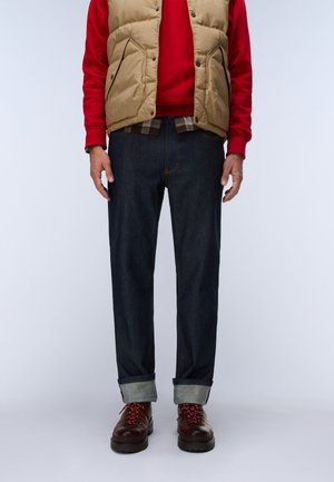 Man wearing beige puffer vest, red sweatshirt, dark cuffed jeans, and brown boots standing against plain background.