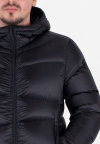 Doudoune noire avec capuche, dotée d'une fermeture éclair et d'un motif matelassé. Le tissu est lisse et brillant, avec un logo circulaire sur la manche.