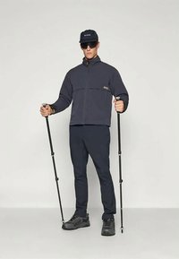 Tumed sinine tõmblukk-kaelusega jakk tekstuuriga paneelidega, sobivad püksid ja tugev must matkasaapad. Hoidmas matkakeppe, kannab päevitusklaase ja mütsi.