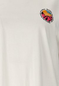 Weißer Stoff mit einem kleinen runden Motiv, das rosa Berge, eine untergehende Sonne und den Text „Atme die frische Luft, beobachte die Schönheit der Natur“ zeigt.