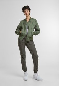 Olivovo zelená bomber bunda s rebrovanými manžetami a pásom, so zipsom na prednej strane a dvoma bočnými vreckami, nosená cez biely top a skombinovaná s olivovými cargo nohavicami.