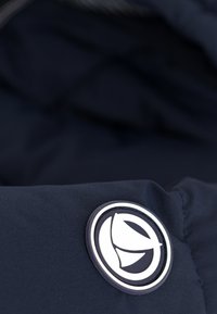 Jūrinis švarkas su apvaliu, guminės tekstūros logotipu, ant kurio pavaizduota lapo dizainas. Lygus audinio paviršius; arti pažvelgus pabrėžiamas logotipo detalumas.