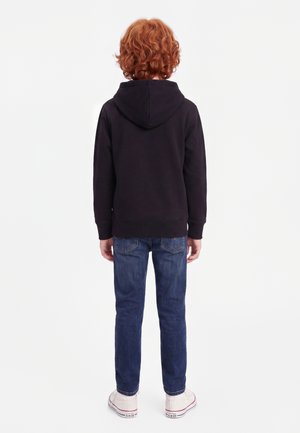 Enfant aux cheveux roux bouclés portant un sweat à capuche noir, un jean bleu et des baskets blanches, debout de dos sur un fond blanc uni.