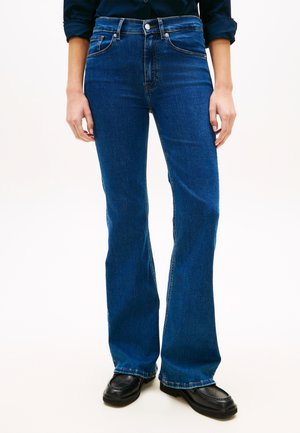 Osoba ve tmavě modrých rozšířených džínách a černých mokasínech stojící s uvolněnými rukama podél těla na jednoduchém pozadí.