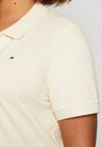Polo de color crema claro con cuello clásico. Cuenta con mangas cortas y un pequeño logotipo bordado en el lado izquierdo del pecho. Textura suave.