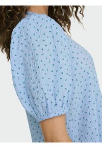 Vaaleansininen paita puffihihalla, joka sisältää ryppyisen tekstuurin ja pieniä vihreitä kukkakuvioita koko ajan. Elastiset hihansuut.