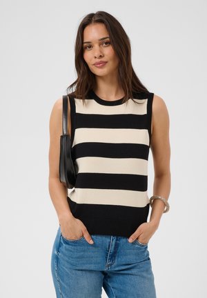 Femme aux cheveux châtain mi-longs portant un haut sans manches rayé noir et beige, un jean bleu et un sac d'épaule noir.