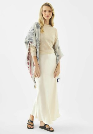 Vrouw draagt een beige trui, witte lange rok, bloemige sjaal losjes over de schouders en zwarte sandalen, staande tegen een effen witte achtergrond.