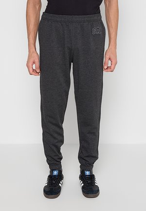 Homme portant un pantalon de survêtement GAP gris foncé et des baskets Adidas noires, debout devant un fond blanc uni.