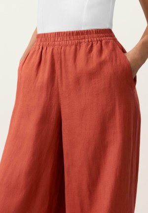 Person trägt lockere Leinenhosen in Rostfarbe mit elastischem Bund und Händen in den Taschen, kombiniert mit einem weißen ärmellosen Oberteil.