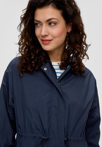 Granatowa lekka kurtka z stójką, ściągaczem w talii i subtelnym połyskiem, z zamkiem błyskawicznym i klamrami.