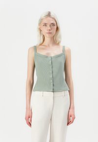 Top in maglia verde a coste con scollatura quadrata, dotato di una fila di bottoni sul davanti. Abbinato a pantaloni chiari e sartoriali.
