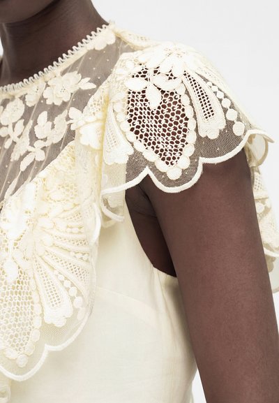 Άτομο με σκούρο δέρμα που φοράει ένα κορυφή σε κρεμ χρώμα με λεπτομερείς λουλουδένιες δαντέλες στους ώμους και την περιοχή του στήθους, σε λευκό φόντο.
