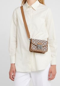 Brązowa skórzana torba na ramię z szarą klapką w wzory, srebrnym okuciem i regulowanym paskiem, noszona na białej koszuli w paski.