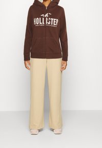 En person som bär en brun zip-up hoodie med texten "Hollister Southern California", beiga vida byxor och ljusrosa sneakers, står.