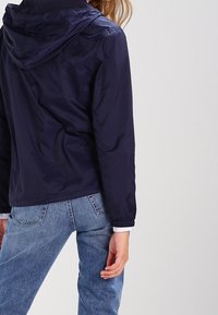 Navy blue waterproof jacket with a hood, elastic cuffs, and a relaxed fit, paired with light-wash denim jeans.