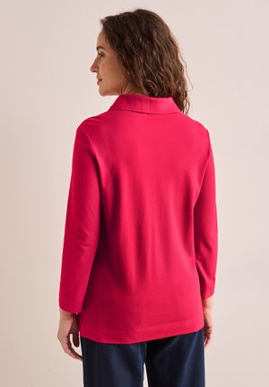 Maglietta rossa a maniche lunghe con colletto, taglio dritto, texture liscia e fessure laterali. Indossata con pantaloni scuri, vista da dietro.