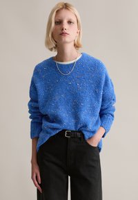 Blauer Strukturpullover mit bunten Sprenkeln, rundem Ausschnitt und überschnittenen Schultern, kombiniert mit hoch taillierter schwarzer Hose und einer silbernen Kettenkette.