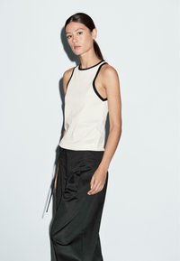 Tank top in cream with black trim, featuring a high neckline and fitted design. Paired with loose-fitting black trousers. Minimalist style.