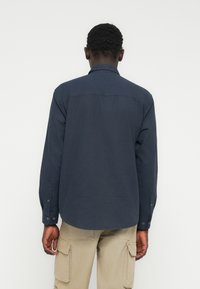 Άνδρας με την πλάτη στραμμένη, φοράει σκούρο μπλε πουκάμισο με μακριά μανίκια και μπεζ παντελόνι καρότσα, μπροστά σε απλό λευκό φόντο.