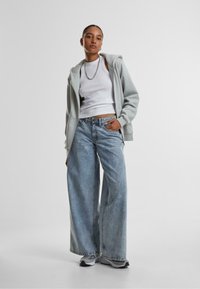 Lys grå hettegenser med glidelås, hvit tanktop og vide lys blå jeans. Sølvkjede som detalj. Joggesko med svart og hvit detaljer.