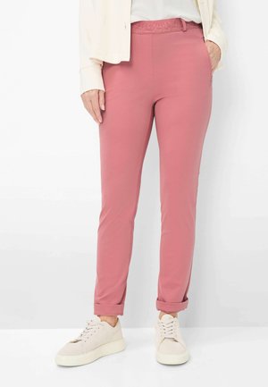 Persona che indossa pantaloni rosa aderenti con risvolti, una giacca bianca e sneakers bianche, in piedi contro uno sfondo semplice.