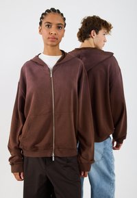Brązowy rozpinany hoodie o luźnym kroju, z dużymi kieszeniami i kapturem. W zestawieniu z luźnymi ciemnymi spodniami oraz jasnoniebieskimi dżinsami w tle.
