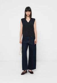 Donkergrijze mouwloze top met een V-hals, gecombineerd met wijde, donkerblauwe denim jeans. Het model houdt een kleine witte handtas vast en draagt bordeauxrode flats.